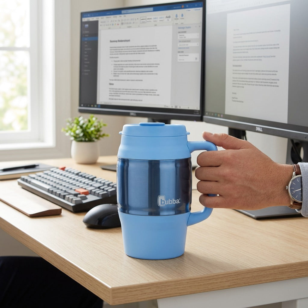 Hand holding a 34 oz bubba Classic Insulated Mug in Daydream blue at an office desk with dual monitors.
